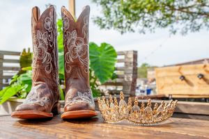 Western boots, tiara, and rose gold accessories arranged on wooden outdoor surface.