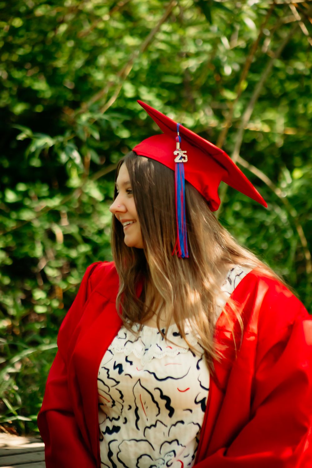 Graduate in red cap and gown smiling while looking to the side against green foliage background.