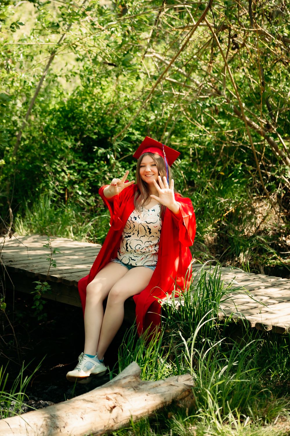 A graduate in a bright red cap and gown sits on a rustic wooden bridge surrounded by lush greenery on a sunny day.