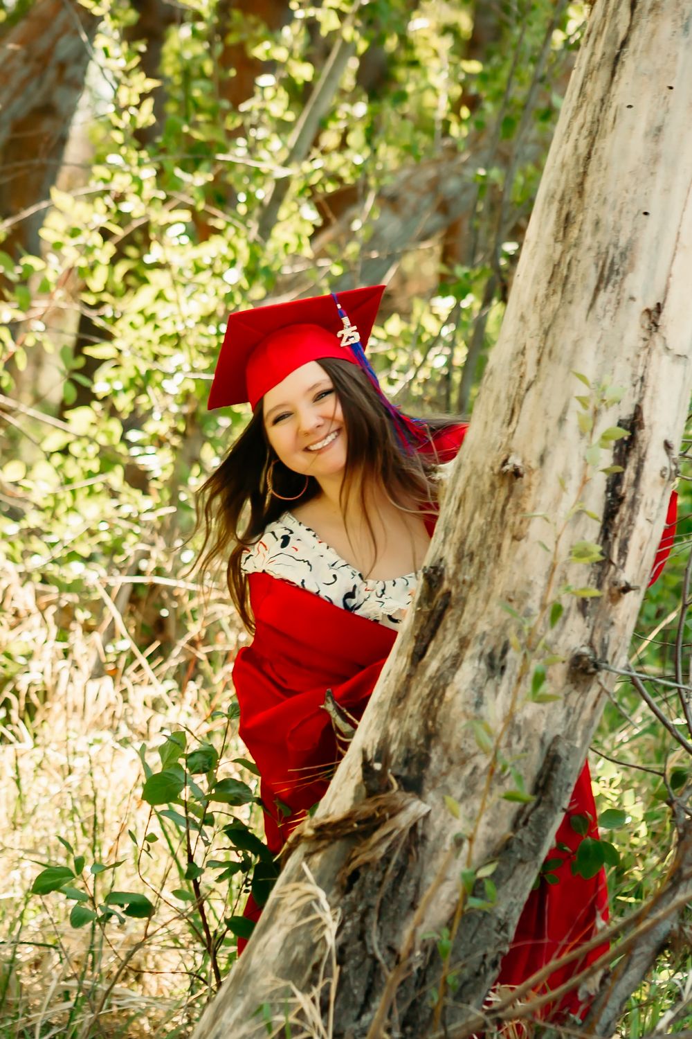 A graduate in a bright red commencement cap and gown poses playfully among trees in a sunlit outdoor setting.