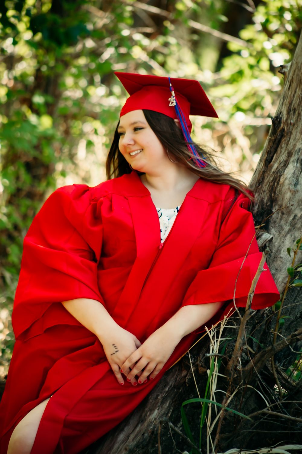 A graduate in a bright red cap and gown sits outdoors against a tree trunk in a natural setting.