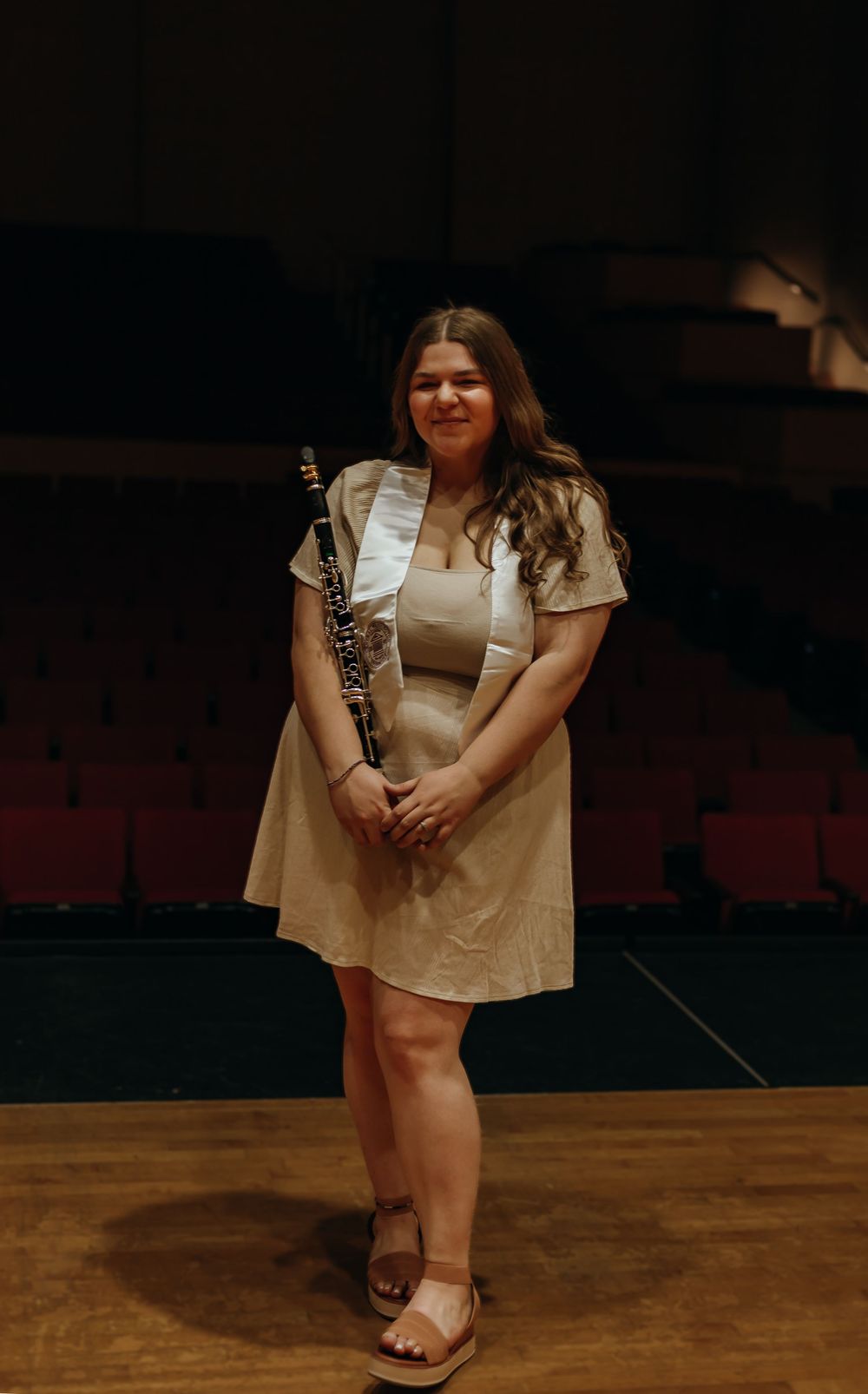 Musician in a beige dress stands on stage holding a clarinet in a dimly lit auditorium.