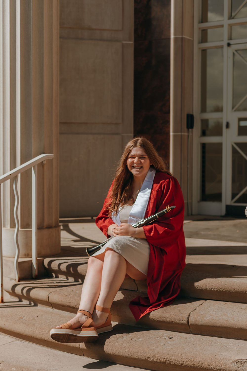 Musician in red cardigan sits on outdoor steps with musical instrument in sunlit setting.