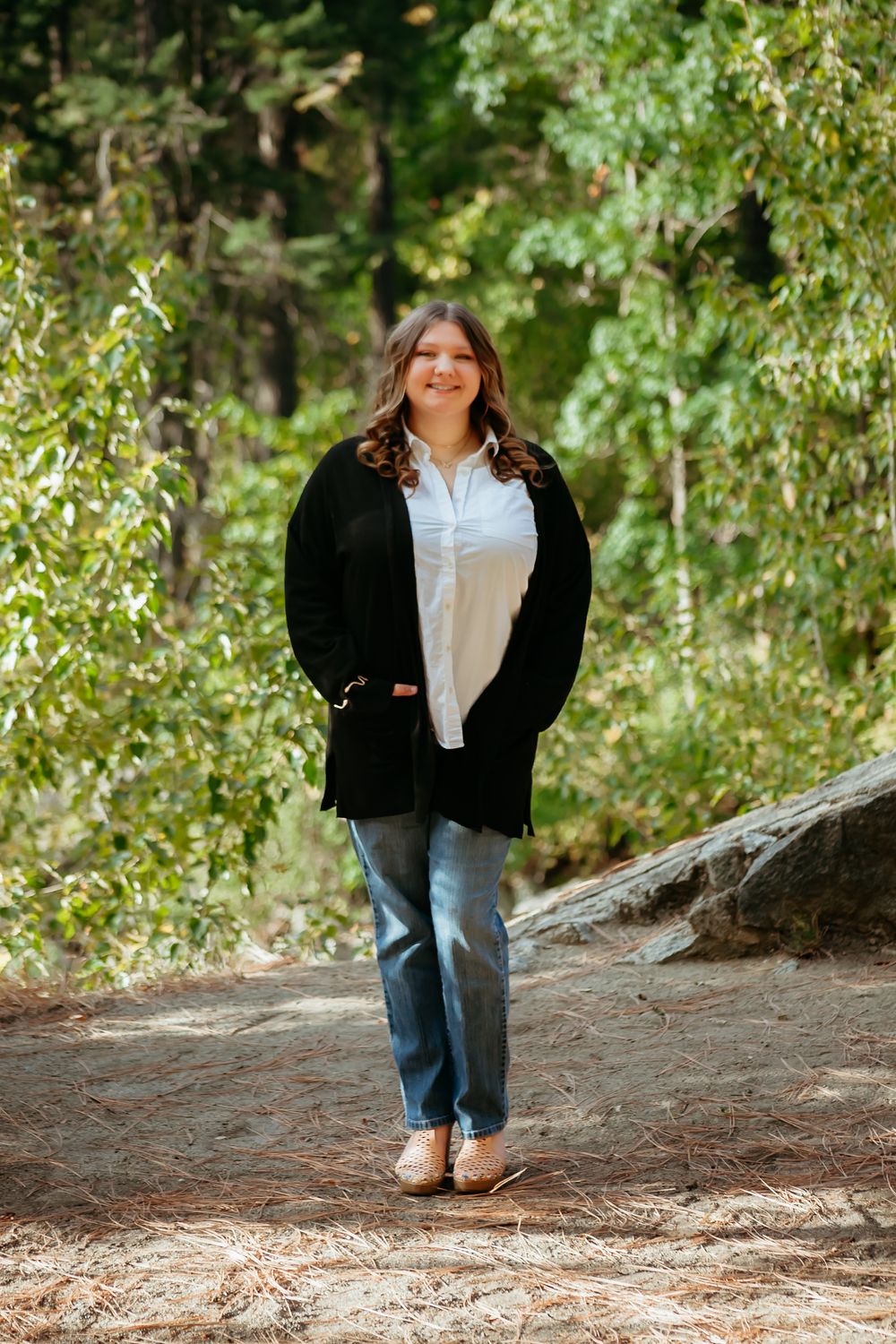 Someone wearing a black cardigan and jeans stands smiling on a dirt path surrounded by green trees in nature.