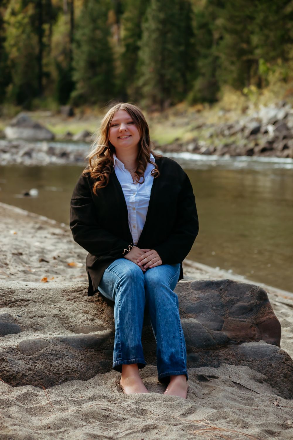 A person wearing a black cardigan and jeans sits relaxed on a large rock by a serene riverside with trees in the background.