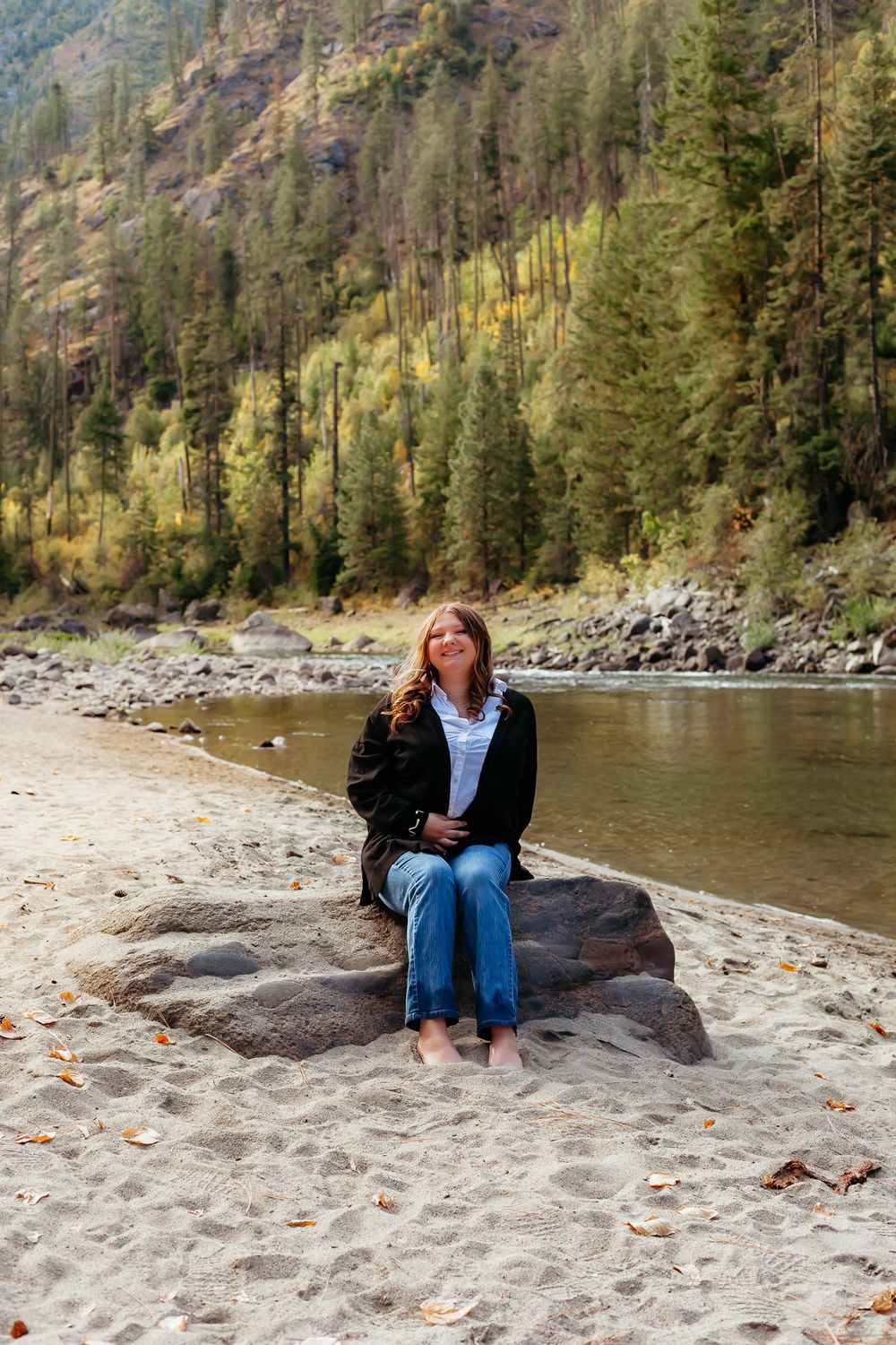 A peaceful riverside beach scene with pine trees on a mountainside and someone relaxing on a large rock near the water.