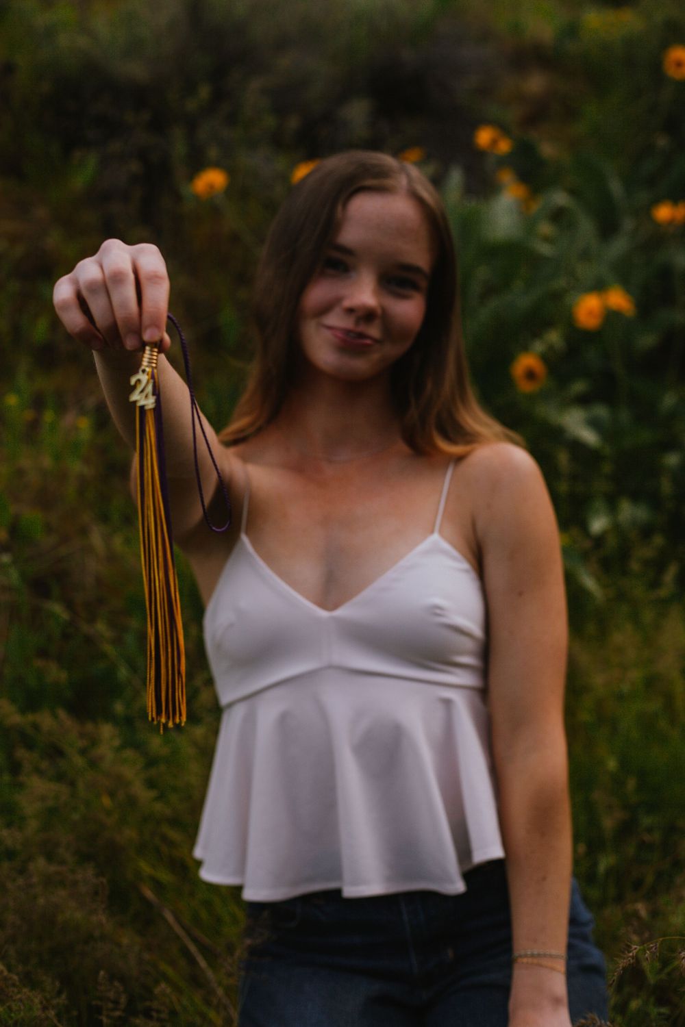 A person in a white top holds up a brown tassel ornament in front of a garden with orange flowers in the background.