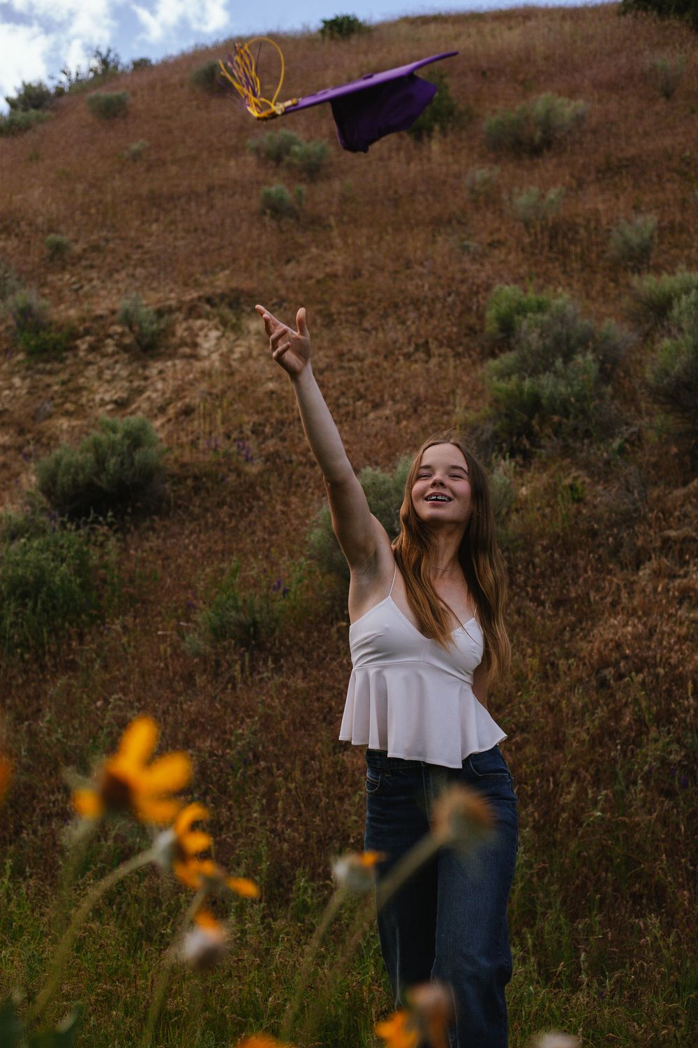 A graduation cap is tossed into the air against a hillside backdrop with yellow wildflowers in the foreground.