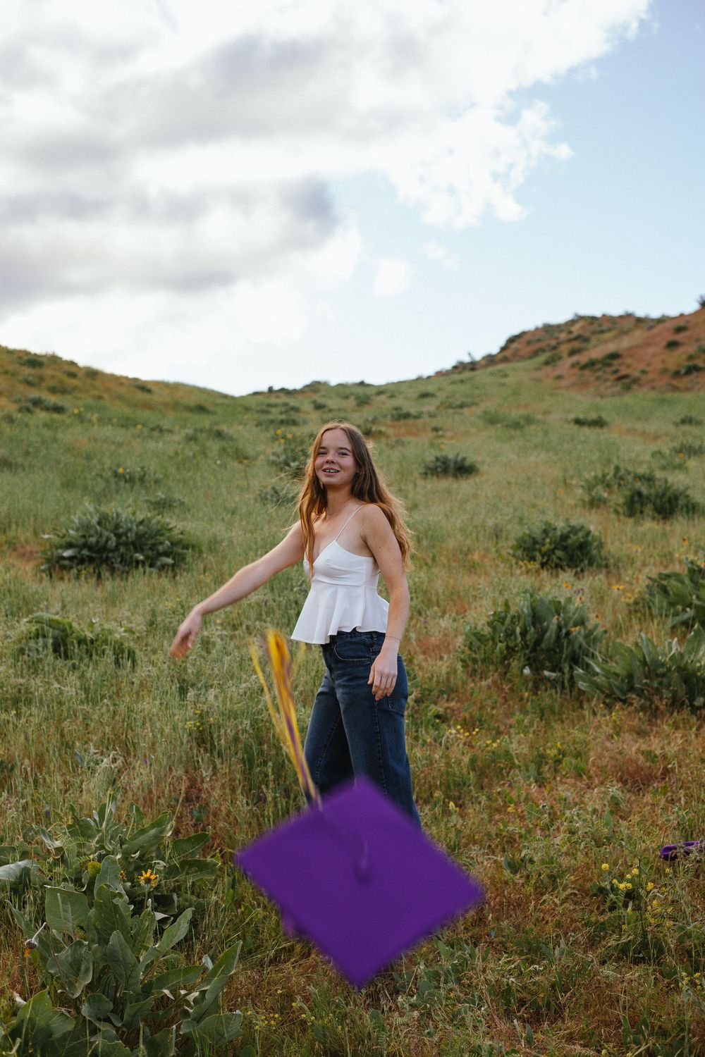 Young woman in white top and jeans holding a purple graduation cap while posing on a grassy hillside.