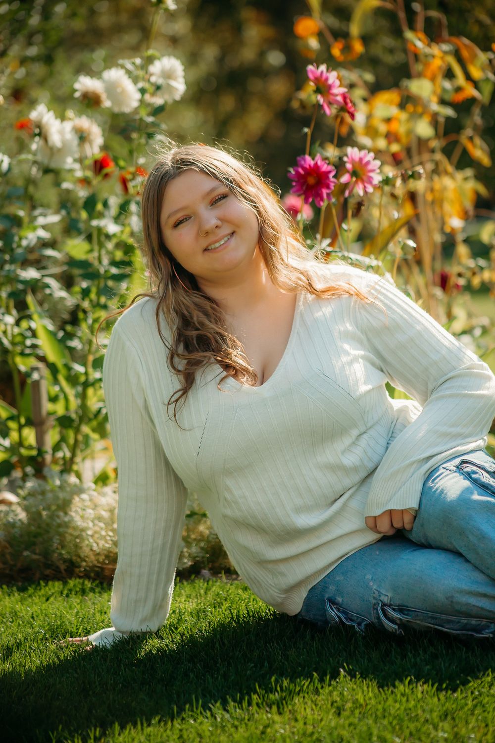A person in a white sweater relaxes on grass surrounded by colorful flowers in a sunlit garden setting.