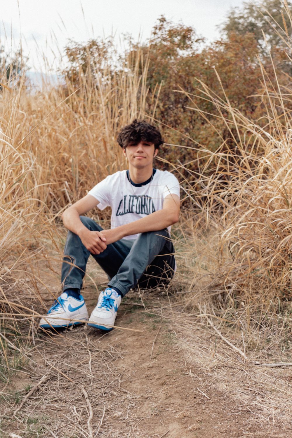 A person sits among tall dried grass wearing a white t-shirt, blue jeans and blue sneakers on a dirt path.