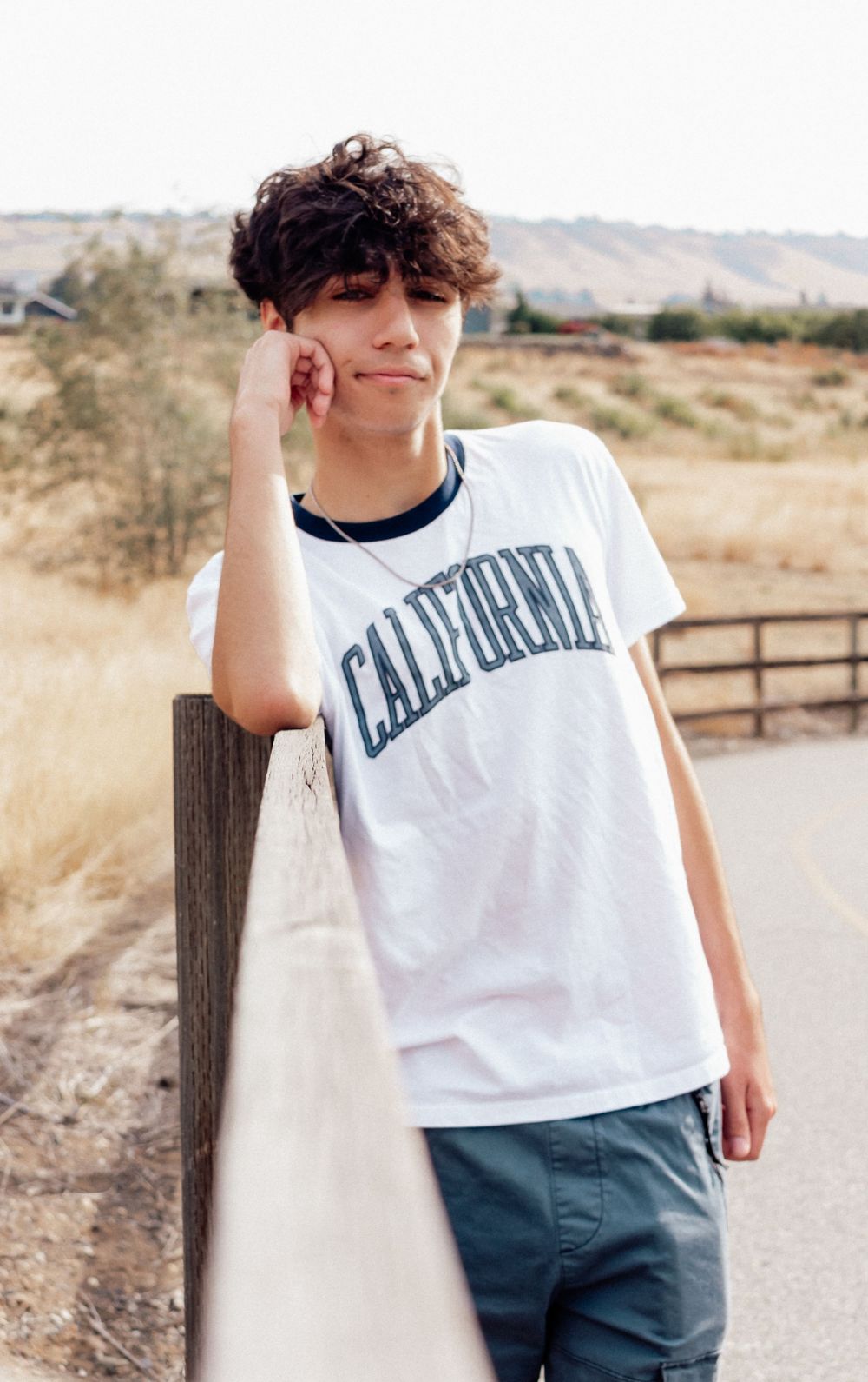 Someone wearing a casual white California t-shirt and blue pants leans against a wooden fence post in a rural setting.