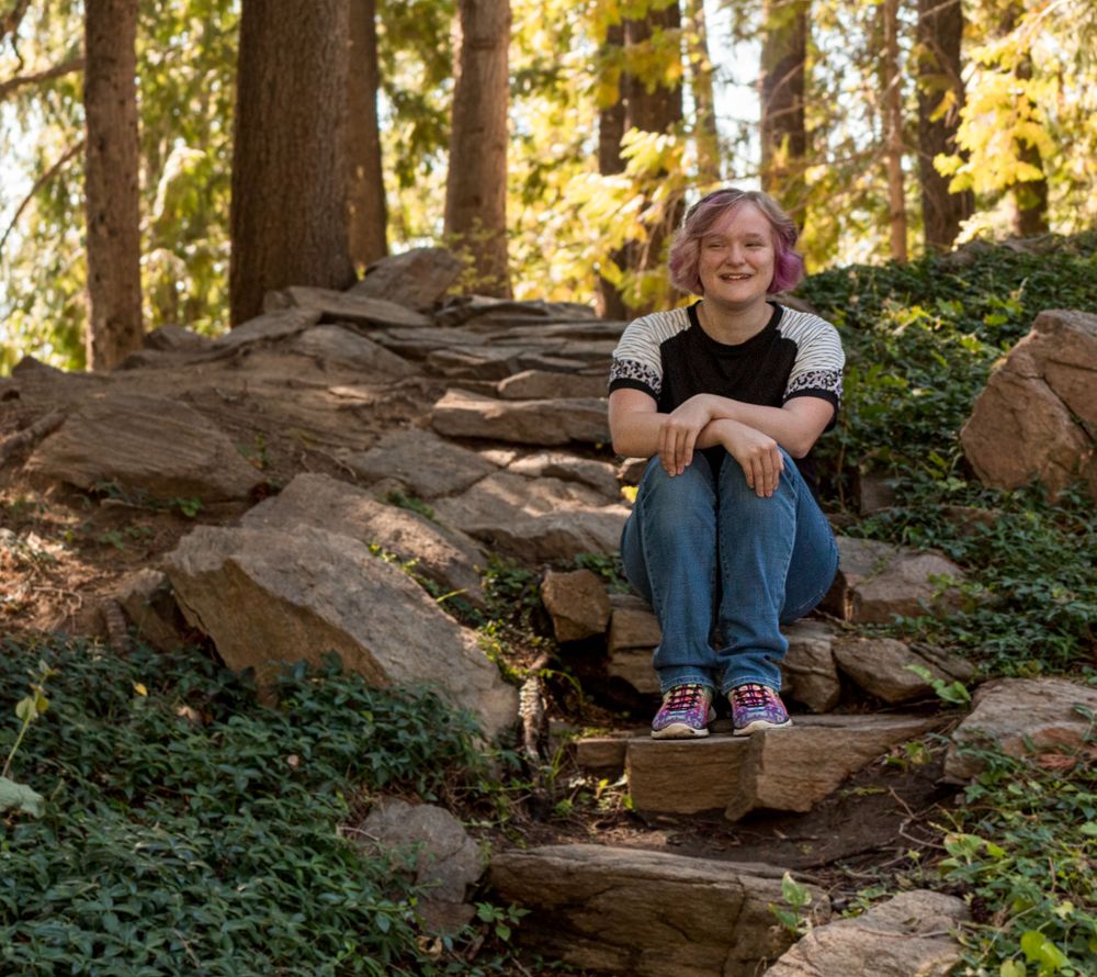 Someone sits casually on natural rock formations in a wooded forest setting with dappled sunlight filtering through trees.