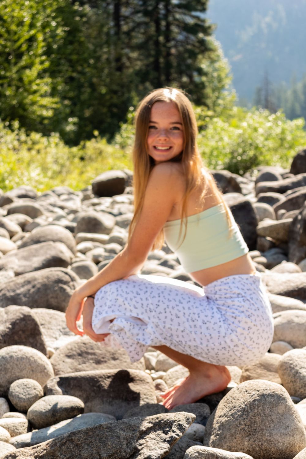 Someone poses on smooth river rocks in a sunny mountain setting wearing patterned pants and a light crop top.