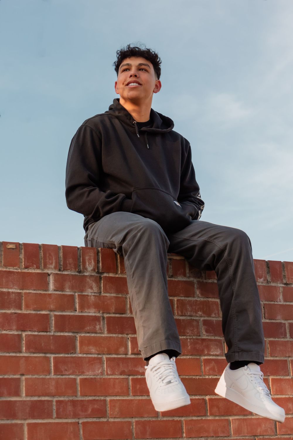 Someone in a black hoodie and gray pants sits casually on a red brick wall wearing white sneakers against a cloudy sky.