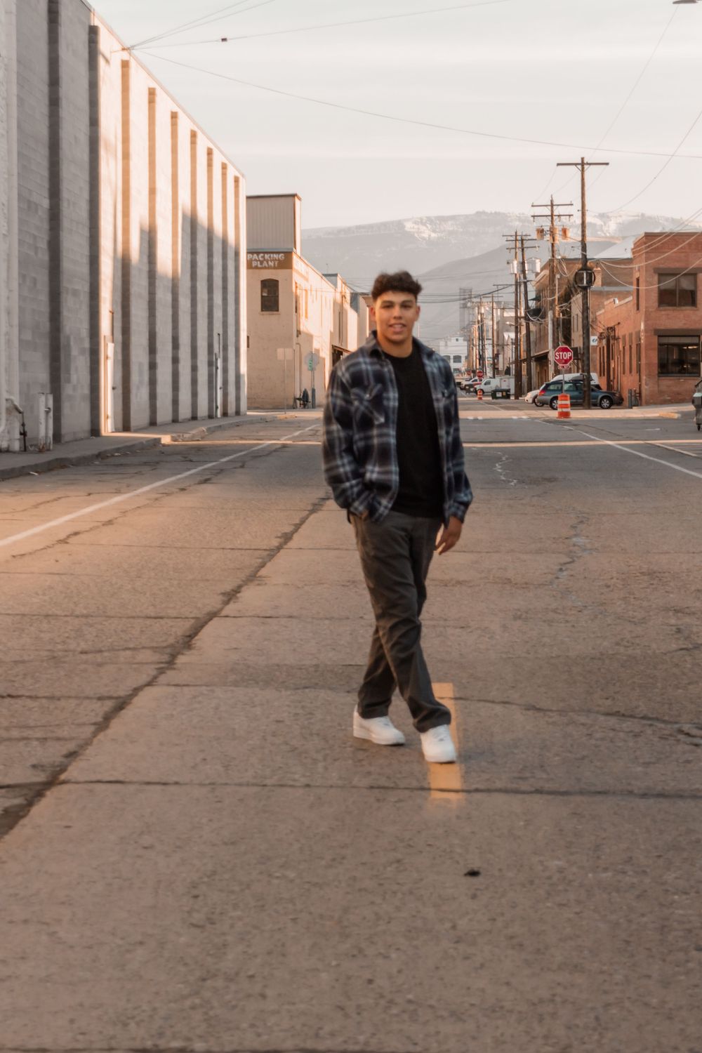 Person in plaid flannel jacket and jeans poses on empty urban street with industrial buildings and mountains in background.