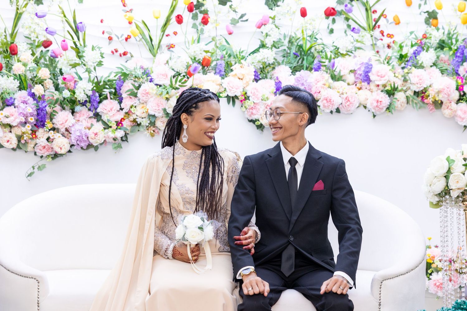 Couple sitting on white couch with floral backdrop sharing intimate moment at their wedding.