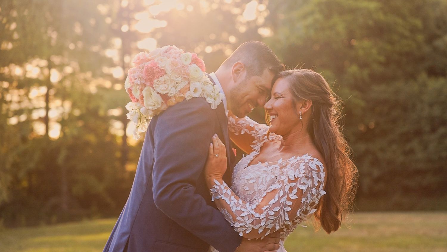 A newlywed couple embraces in a golden sunlit field during sunset while holding a bouquet of pink and white flowers.