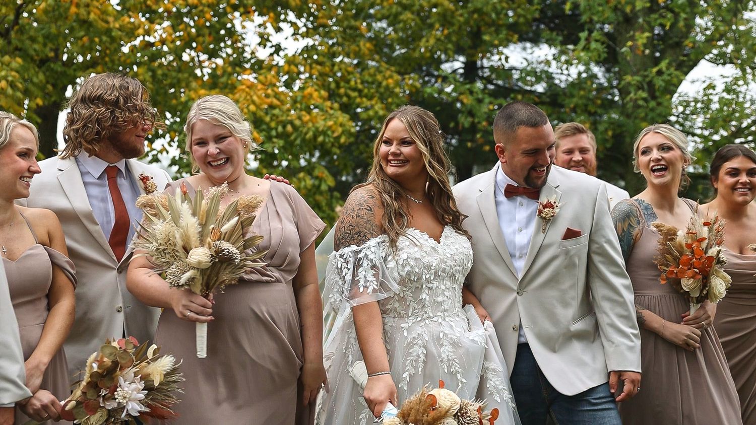 A joyous wedding party wearing beige and white attire walks together outdoors holding bouquets in fall.