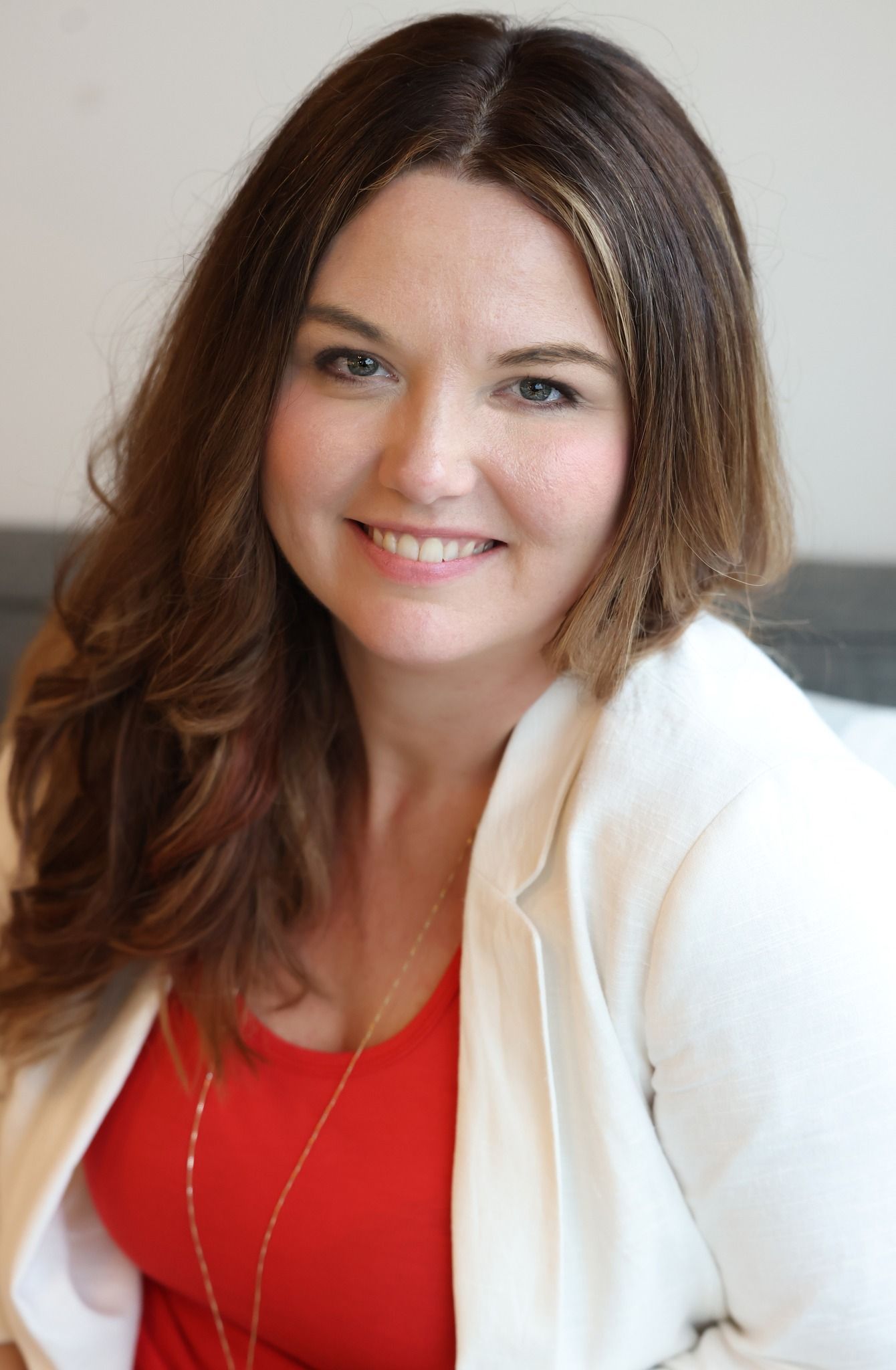 A professional headshot featuring brown hair, a bright red top, and white blazer against a neutral background.
