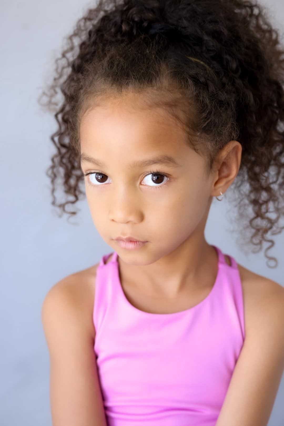 Young child in pink tank top and curly dark hair poses for studio portrait against light background.