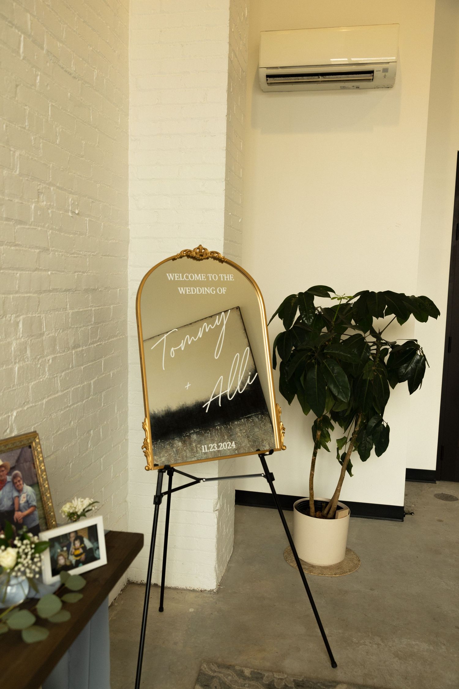 An arched gold mirror welcome sign on an easel next to a potted plant against white brick wall.