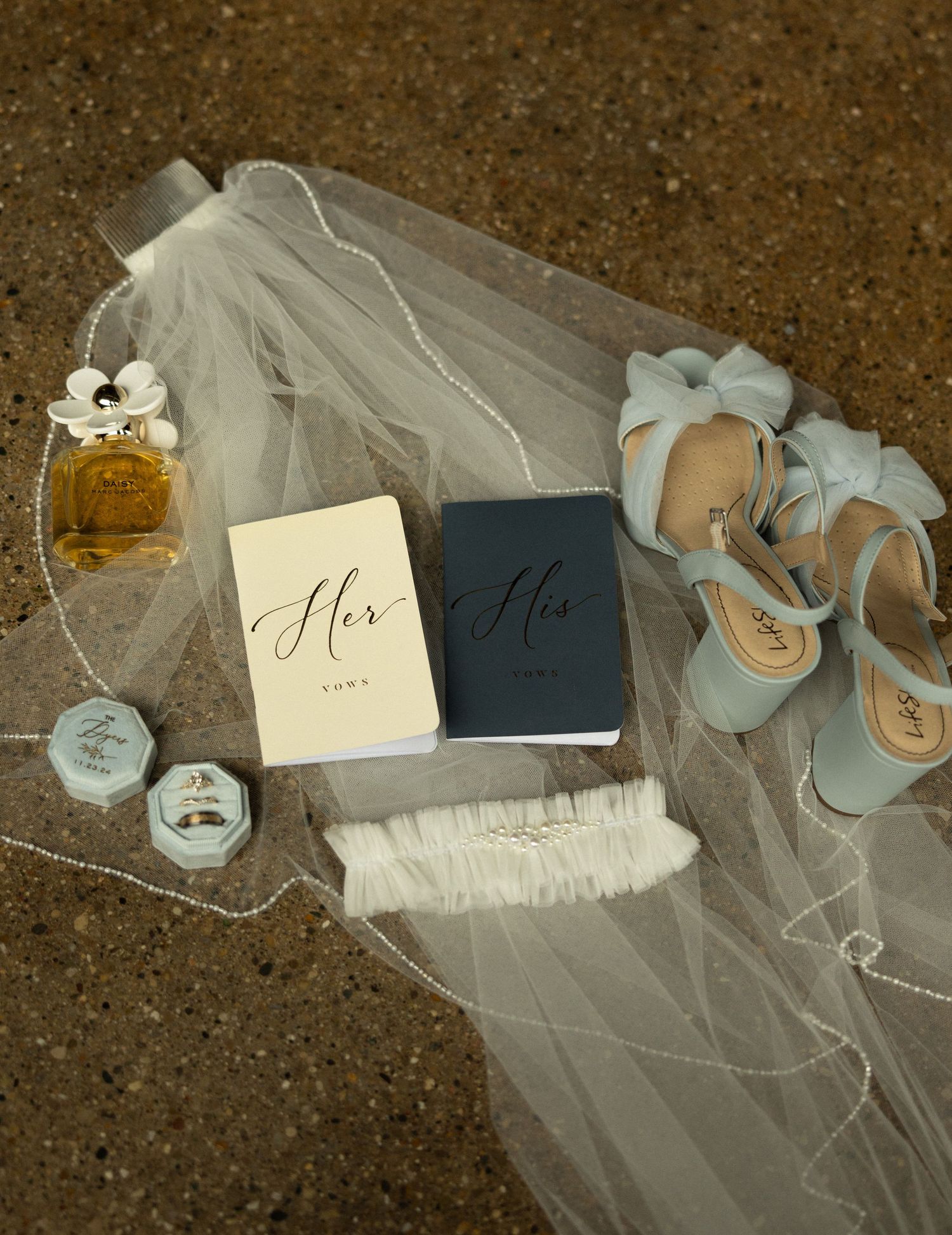 Wedding accessories including veil, shoes, perfume and vow books arranged on concrete surface.
