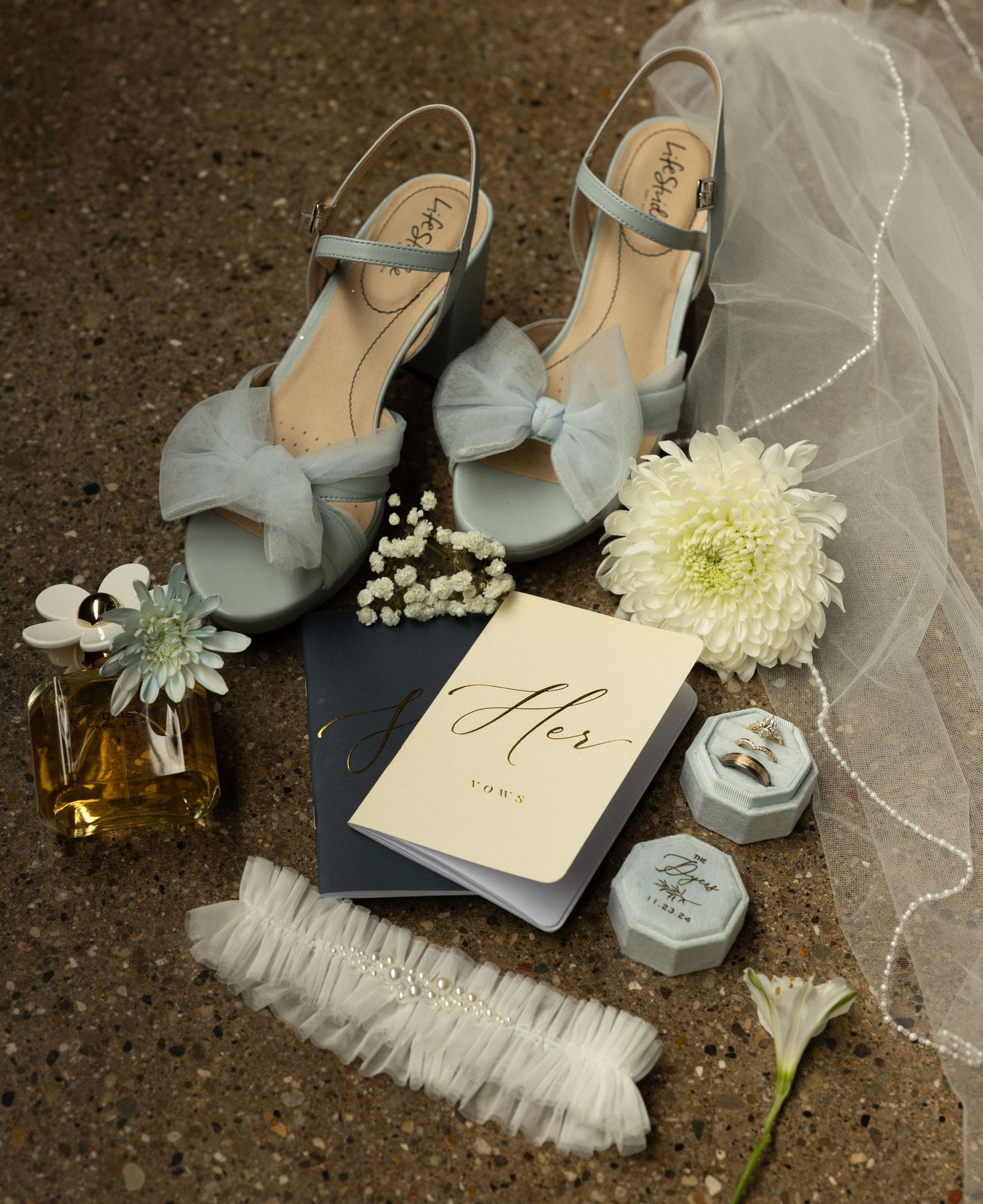 Wedding flat lay with blue bow shoes, veil, flowers and vow books on dark surface.