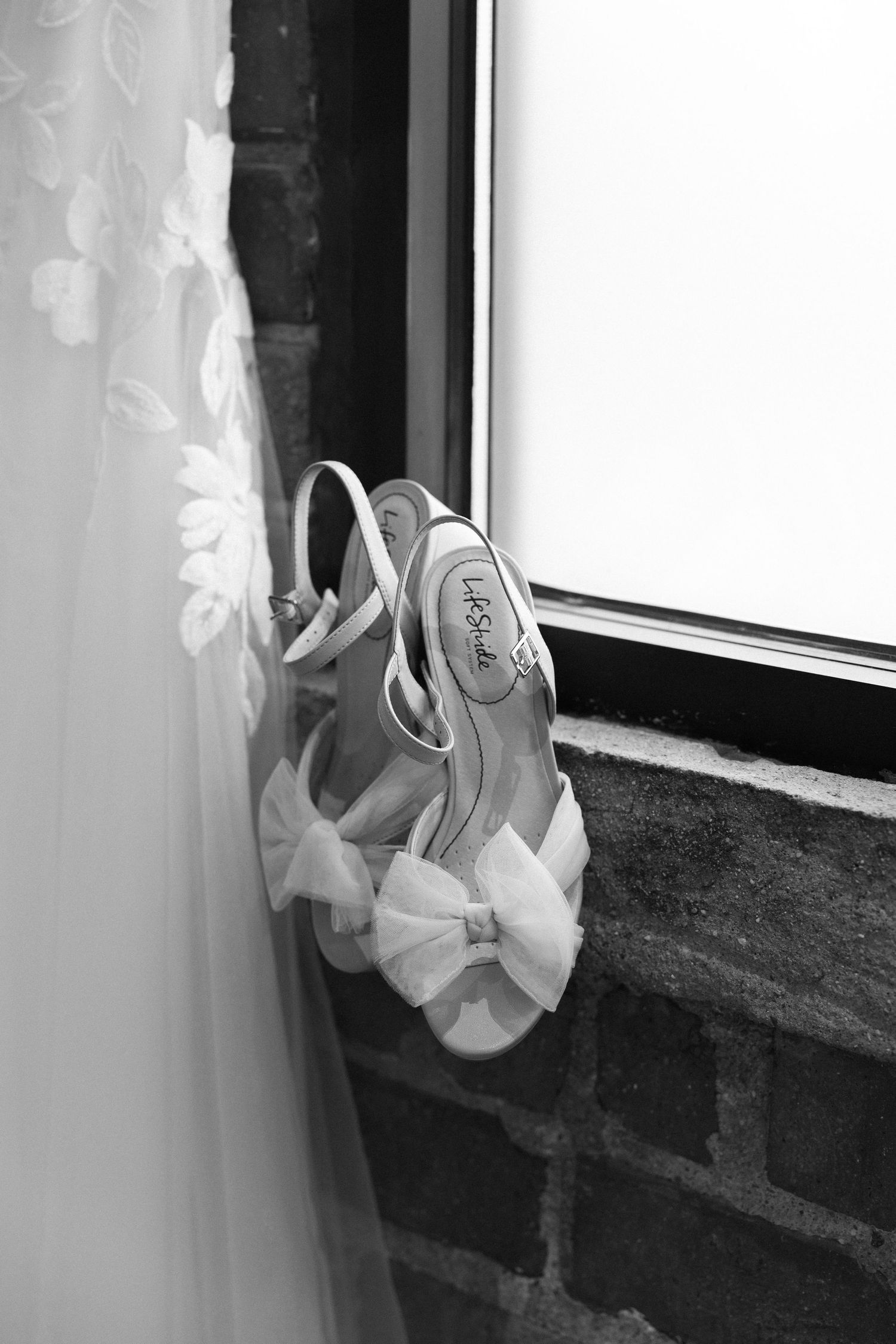Elegant white bridal shoes with bow detail sit on a windowsill in black and white photography.