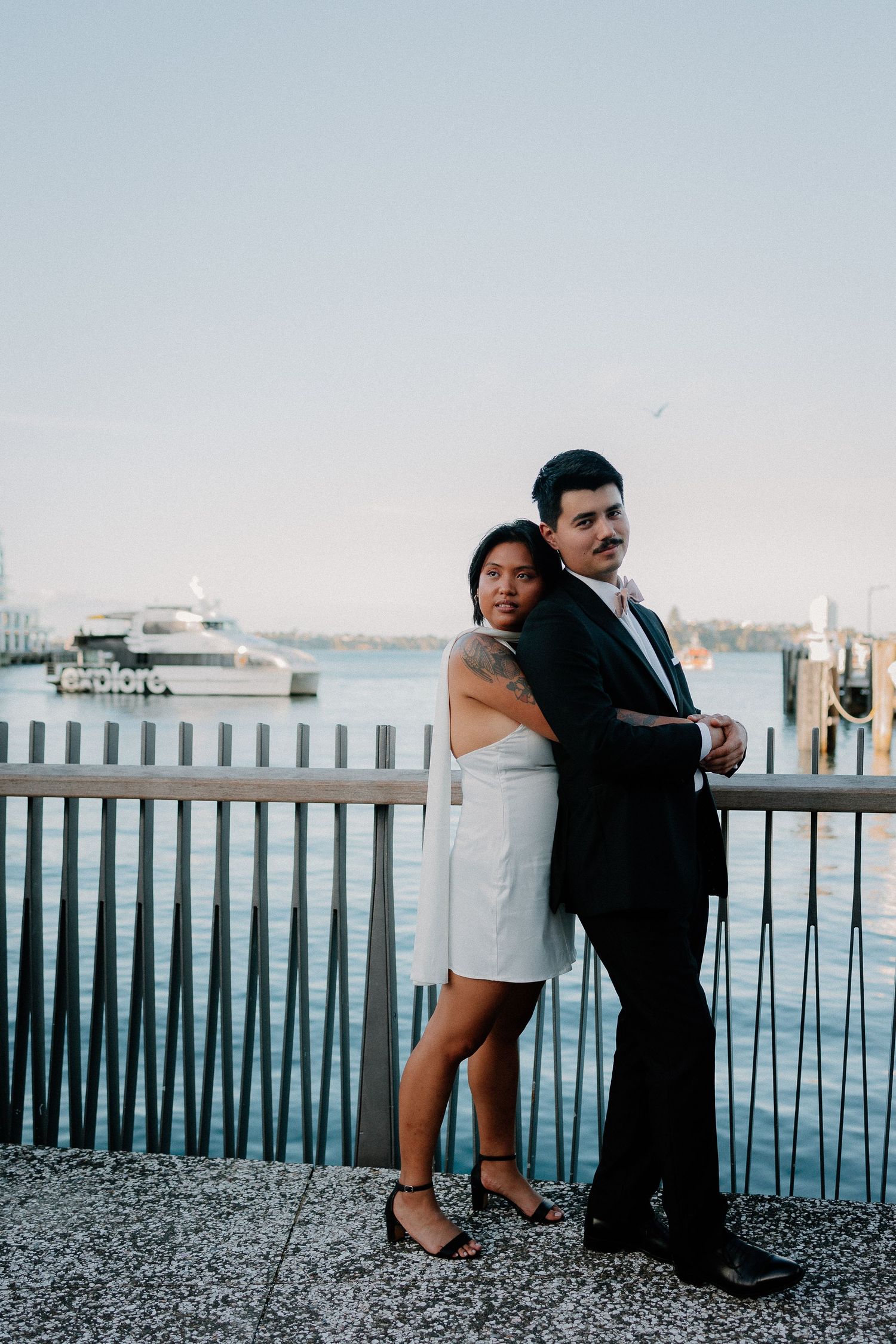 A couple in wedding attire poses together against a railing at a waterfront marina with boats in the background, in Auckland CBD.