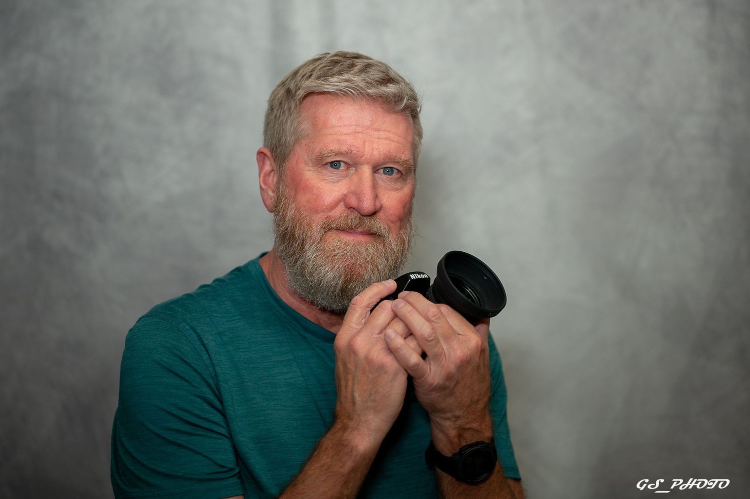 Middle-aged man with gray beard holding a camera, wearing a teal shirt, posing for a portrait photo.