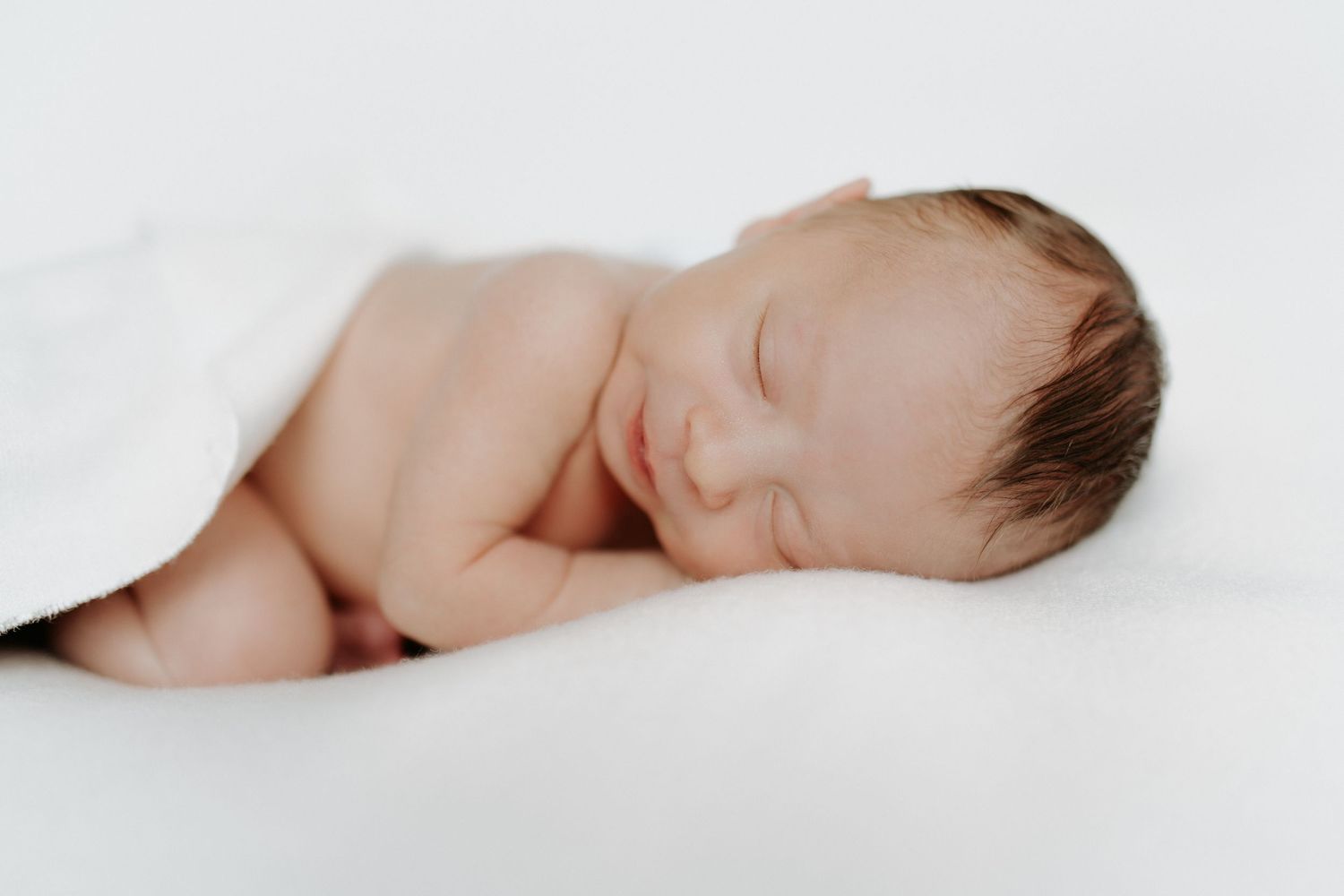 A newborn baby sleeps peacefully on soft white fabric with one tiny arm raised.