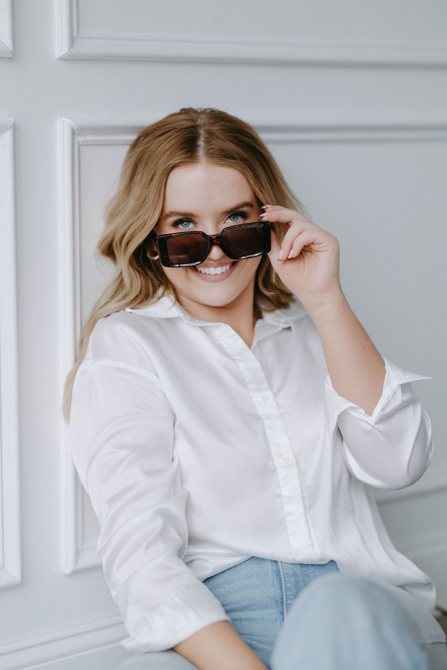 Fashion portrait featuring white button-down shirt and trendy sunglasses against white wall.
