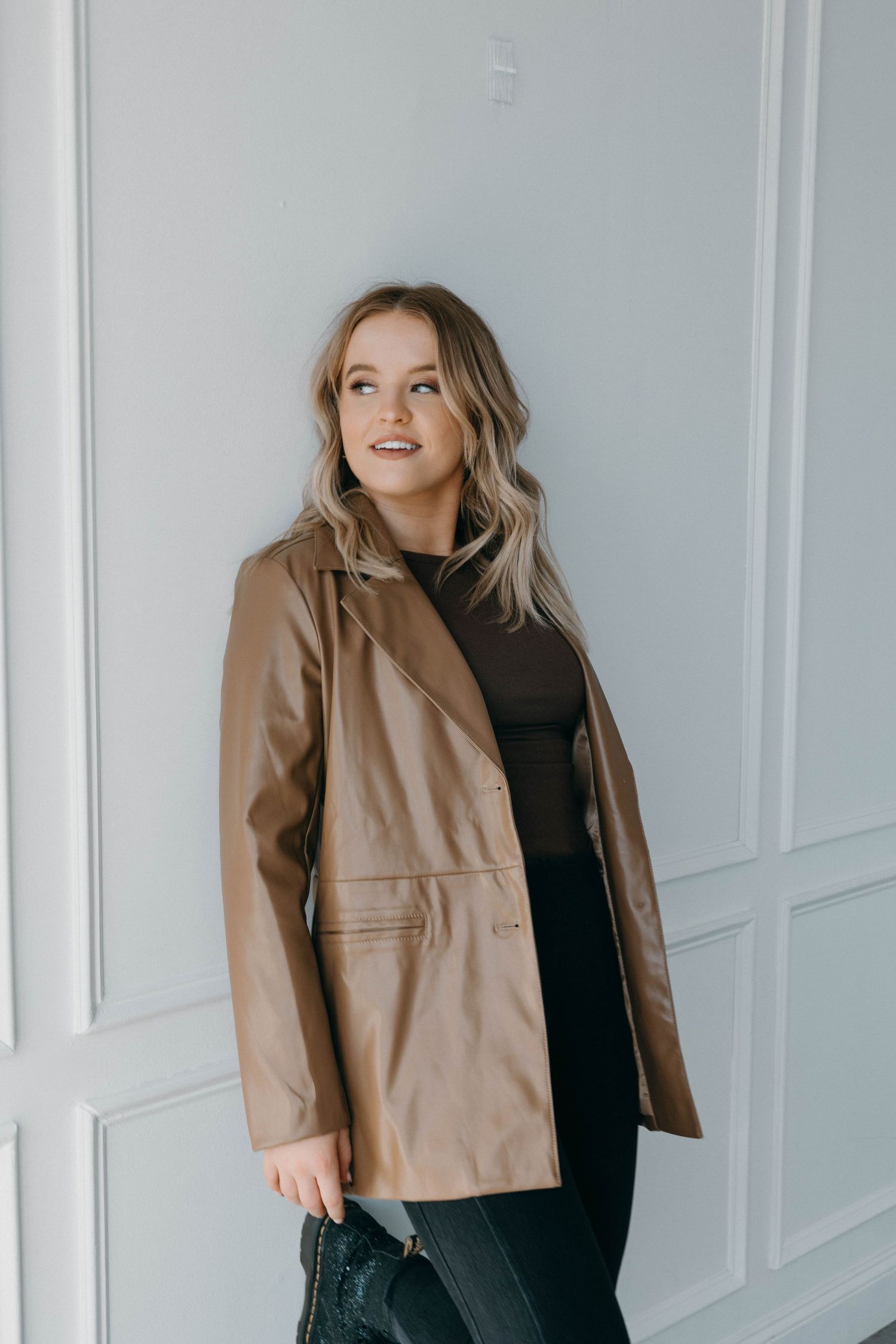 A stylish portrait featuring a tan leather jacket and black outfit against a white paneled wall.