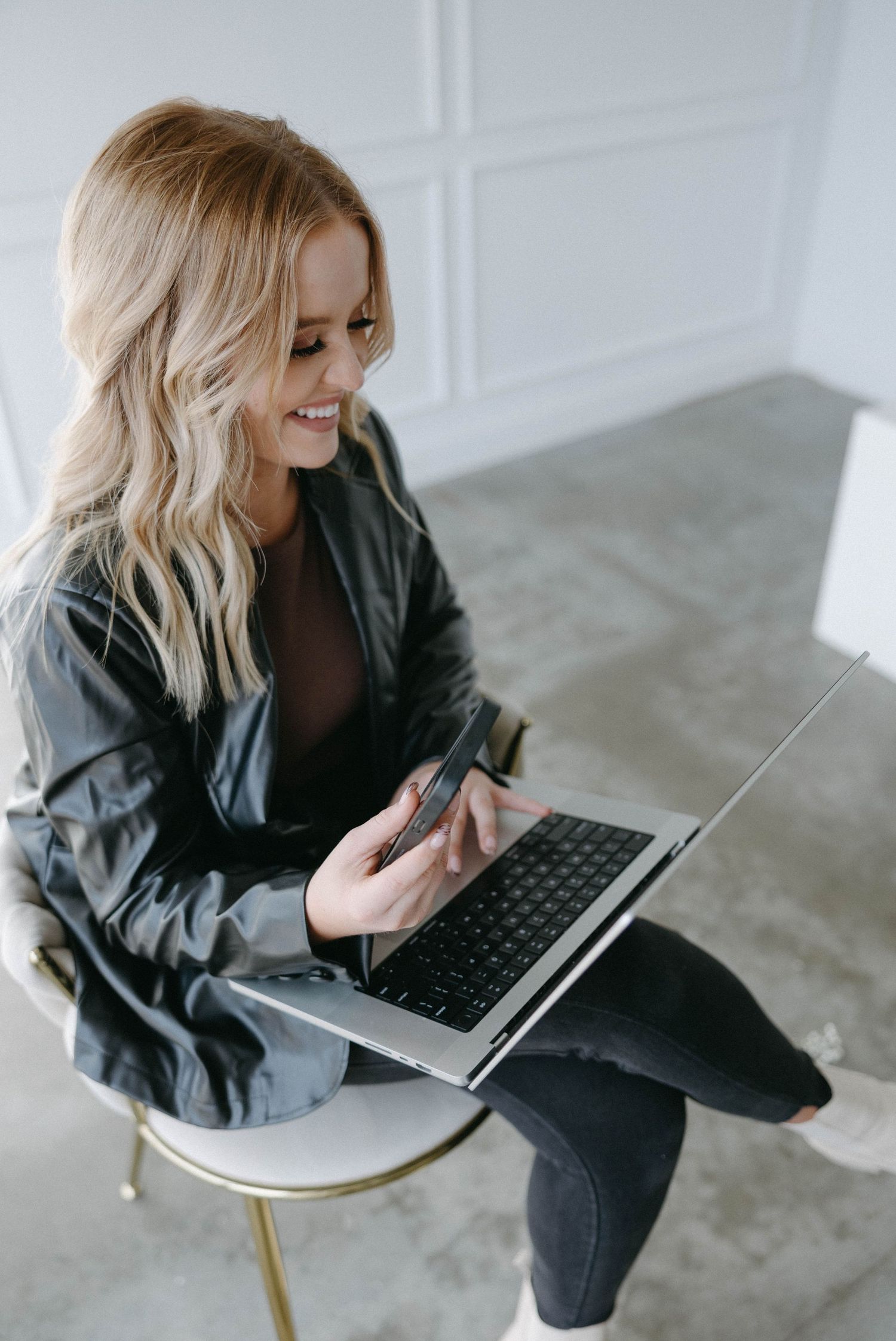 Remote worker in black jacket sits with laptop on modern white chair and smiles while working from home.