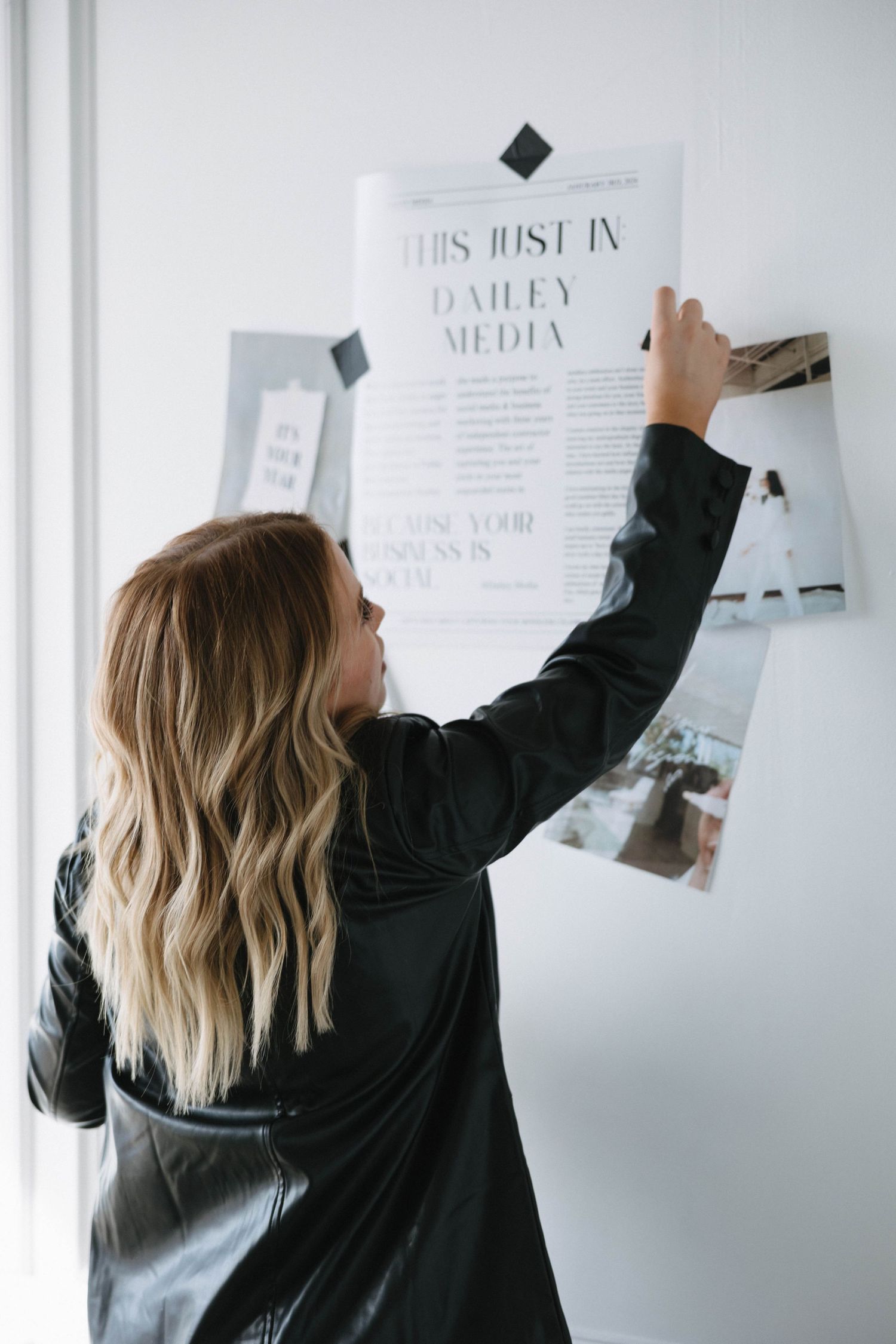 Person in black jacket pins documents to white wall in modern office space.