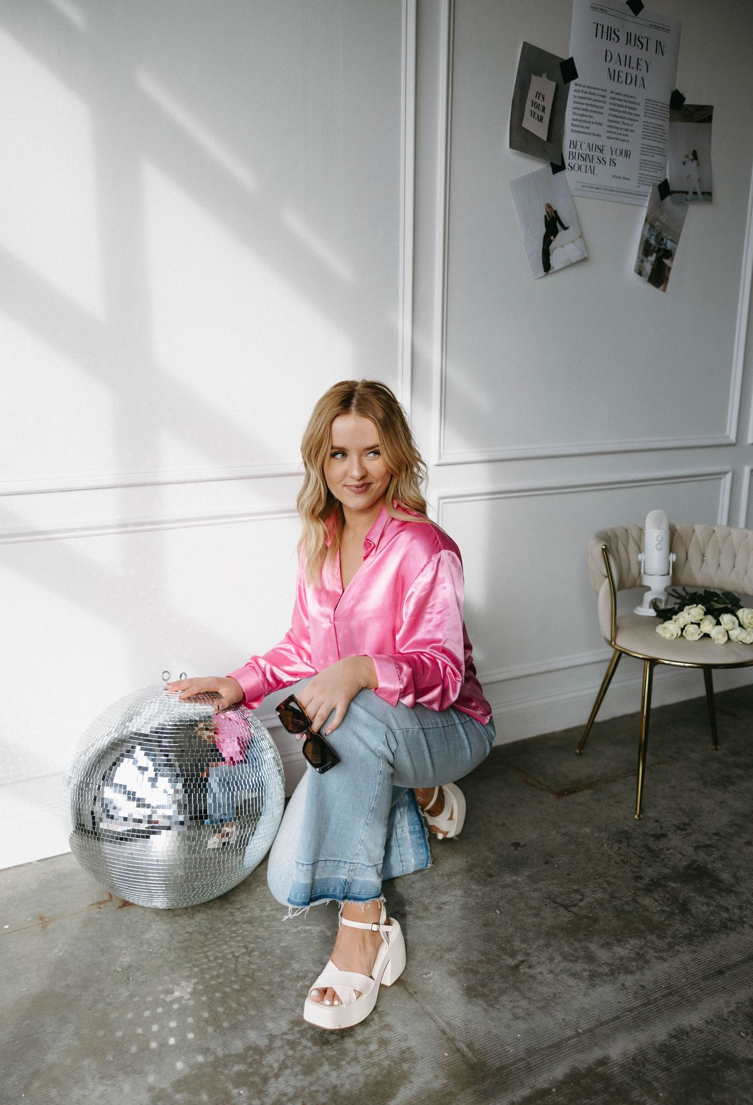 Person in pink satin shirt poses with silver disco ball against white wall.