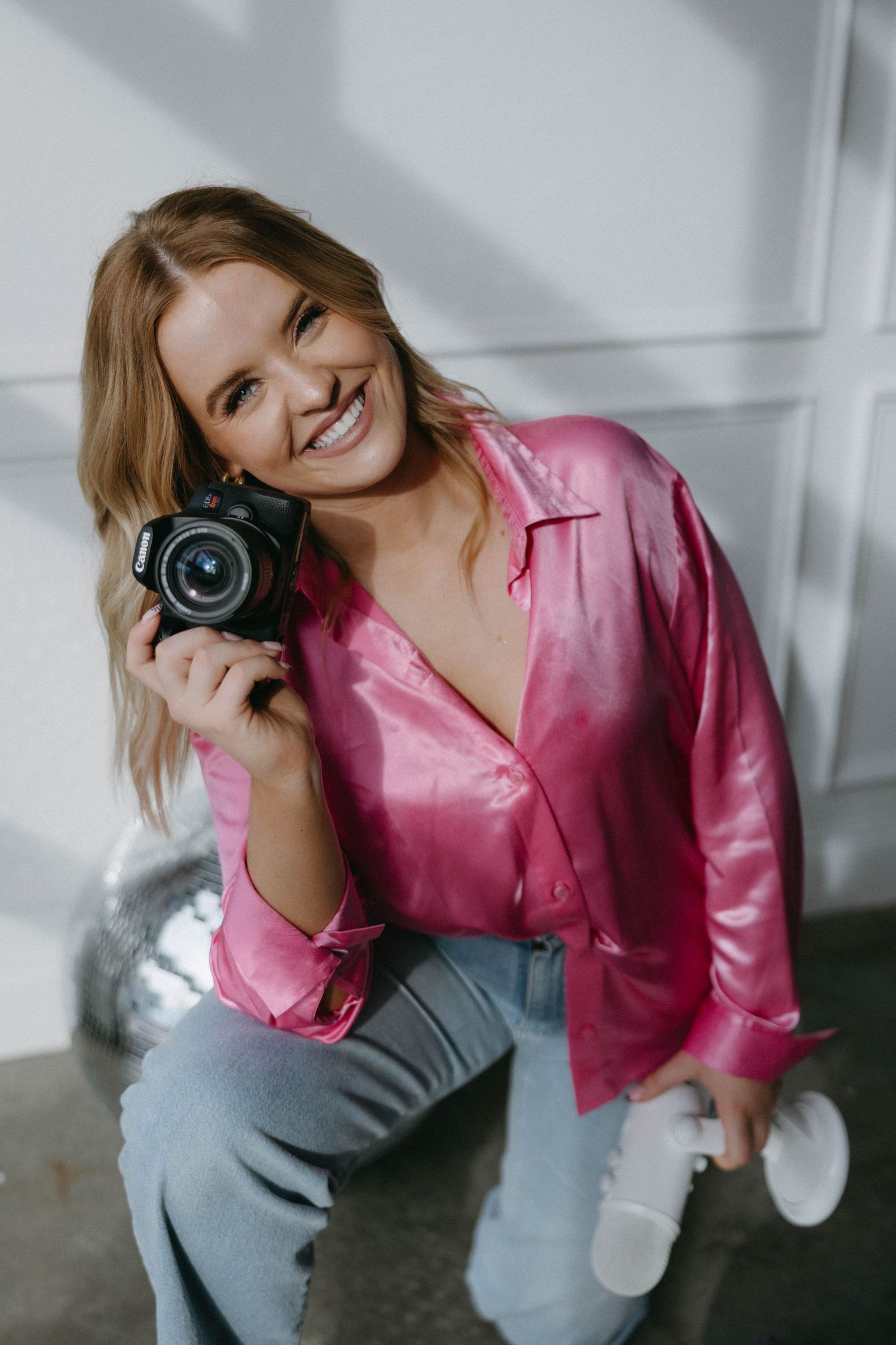 Person in pink shirt holds camera while posing with disco ball in bright room.