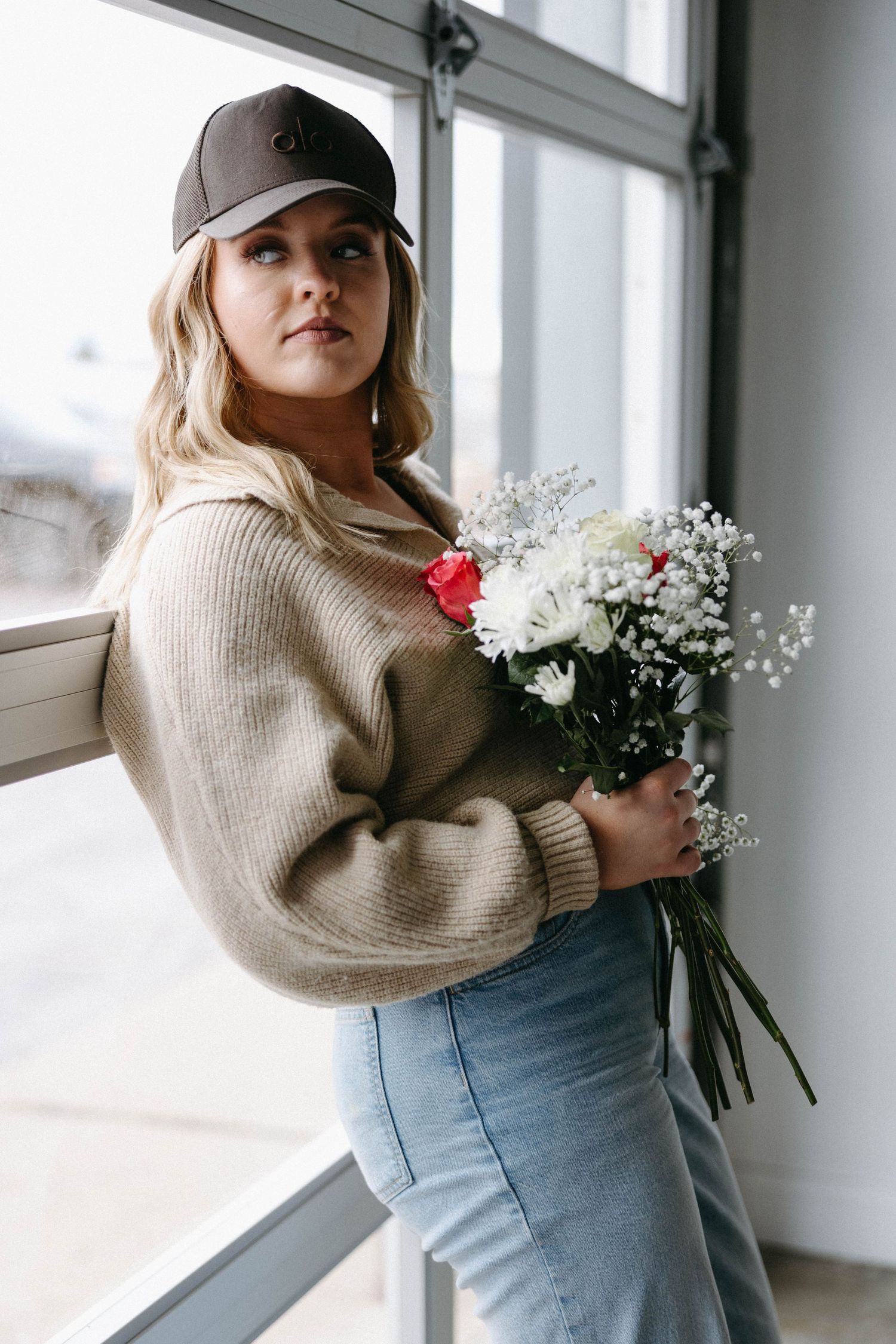 Casual outfit featuring beige sweater and jeans while holding a bouquet of mixed flowers by a window.