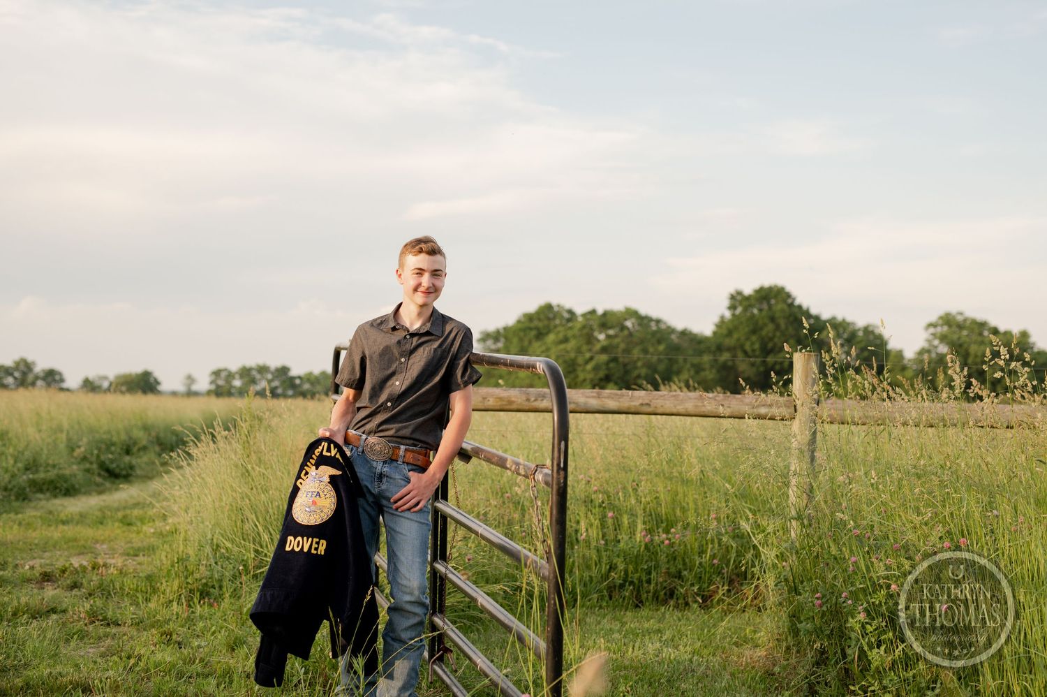Student in casual clothing poses with FFA jacket near farm fence in grassy field.