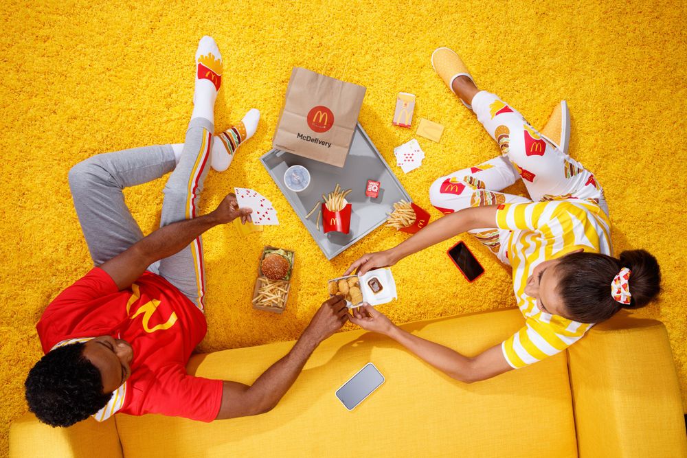 Two people lying on yellow carpet with McDonald's food and drinks scattered around them.