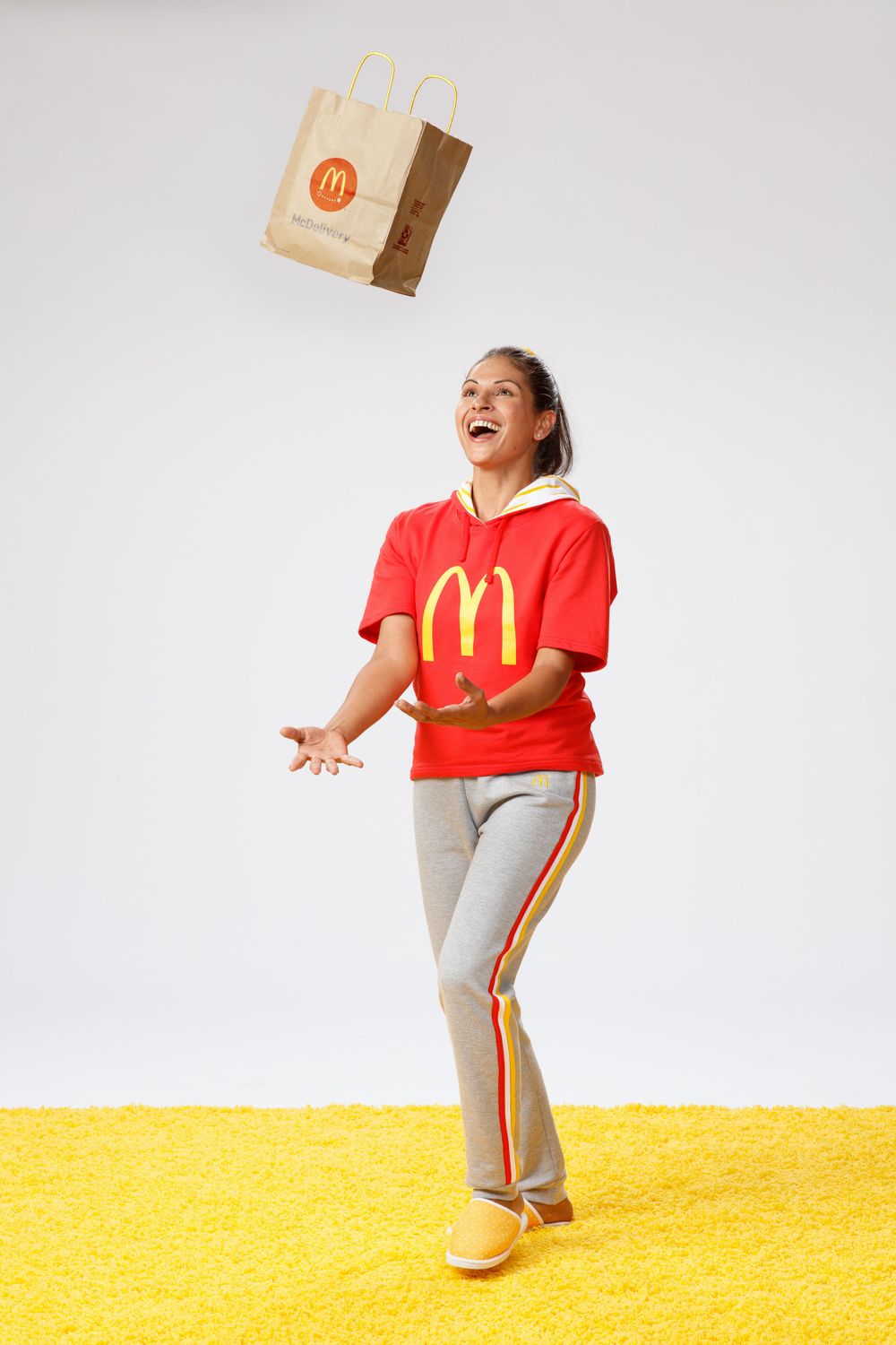 A person in McDonald's uniform stands on yellow background while tossing a paper bag in the air with joyful expression.