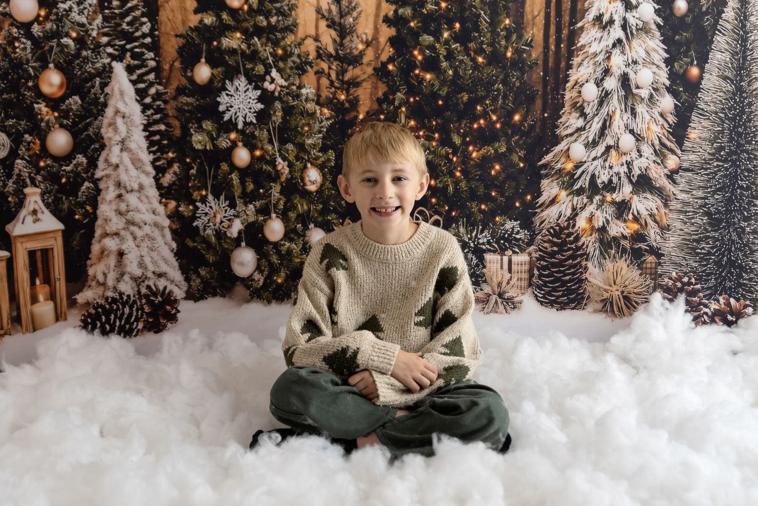 Two children wearing matching sweaters sit cross-legged in front of a whimsical Christmas gingerbread house backdrop.