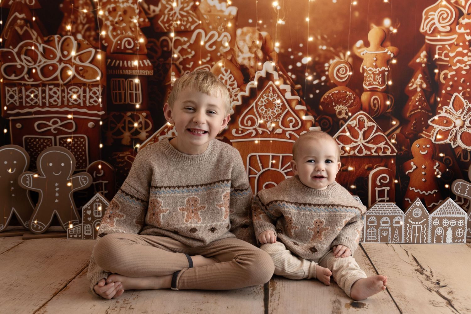 Two children wearing matching sweaters sit cross-legged in front of a whimsical Christmas gingerbread house backdrop.