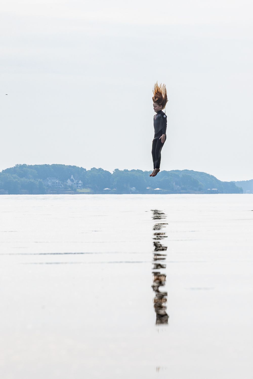 Person jumping high above a reflective water surface with hair flying, trees and overcast sky in the background.