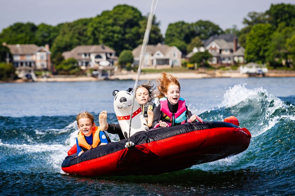 Kids riding a red inflatable tube behind a boat on a lake, laughing and holding a panda stuffed animal.