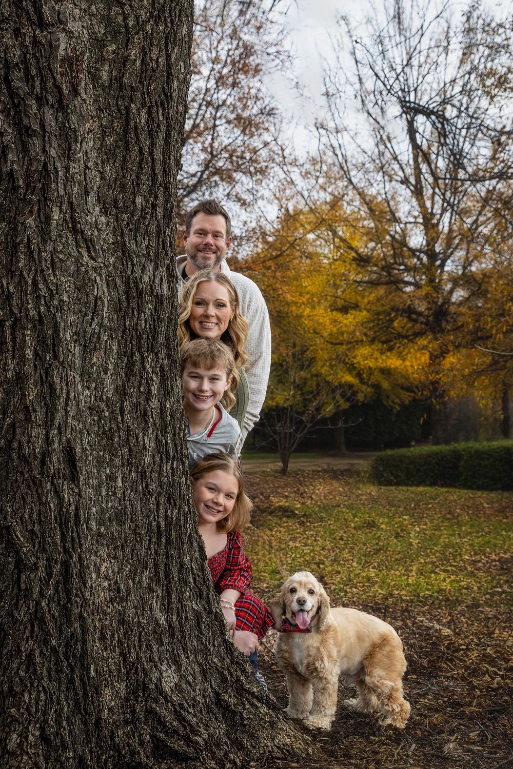 A family gathers around a large tree trunk in a park during fall with their small dog.