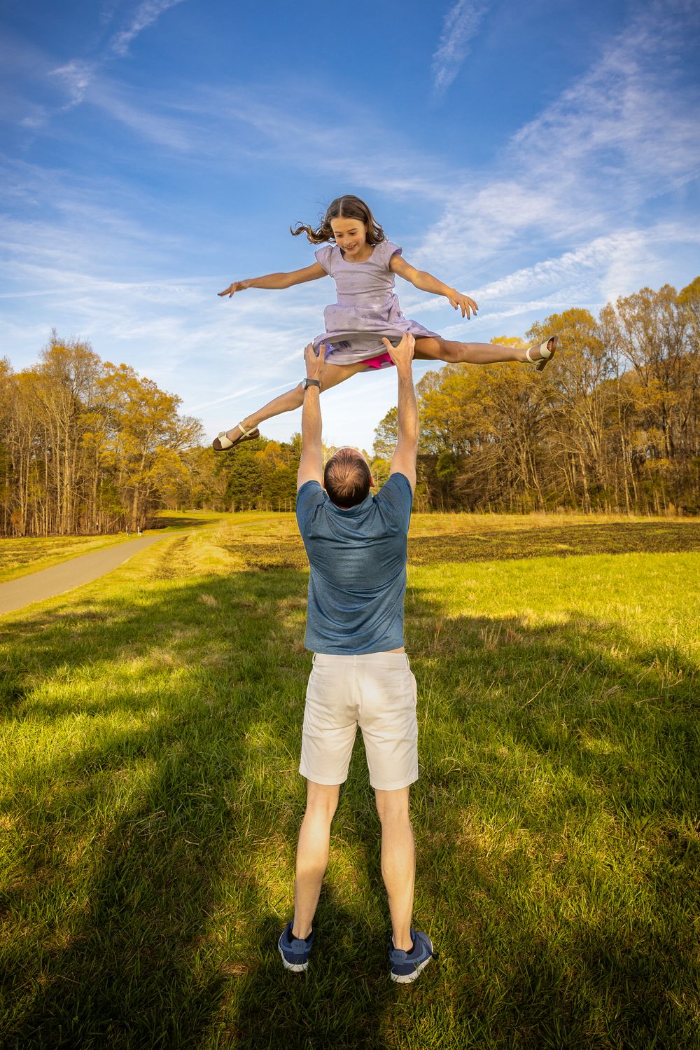 Father lifting daughter high in the air outdoors in a sunny green park, both smiling with joy.