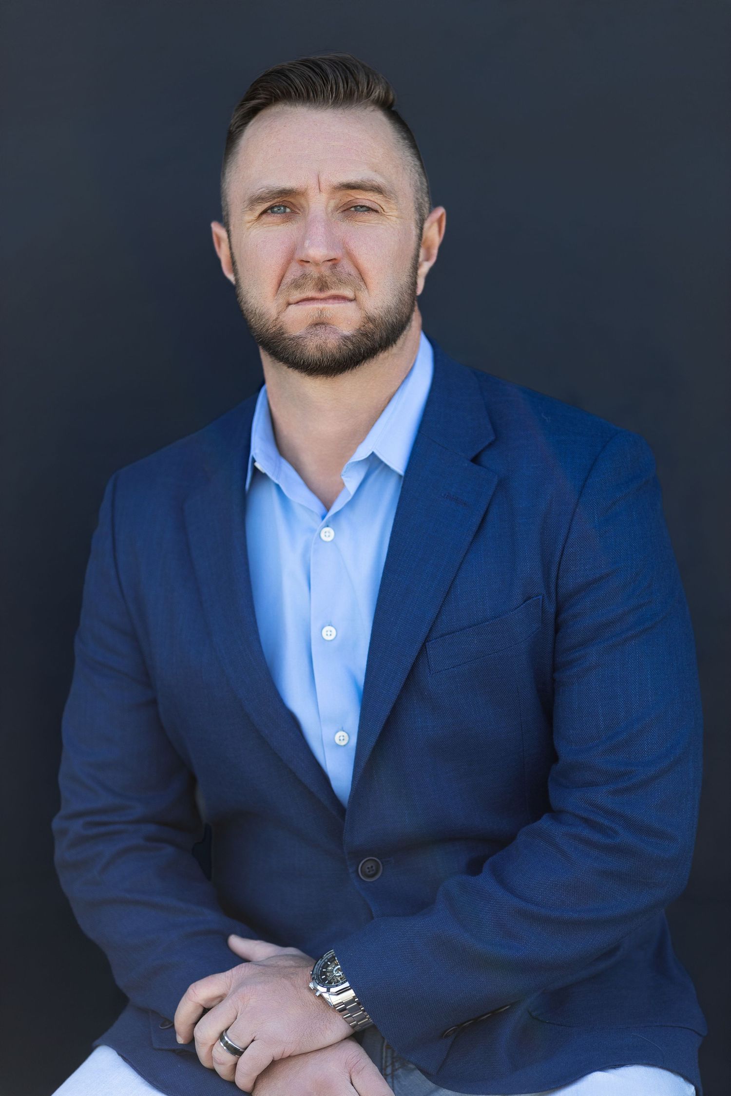 Professional business portrait showing a person in a navy suit and light blue shirt against a dark background.
