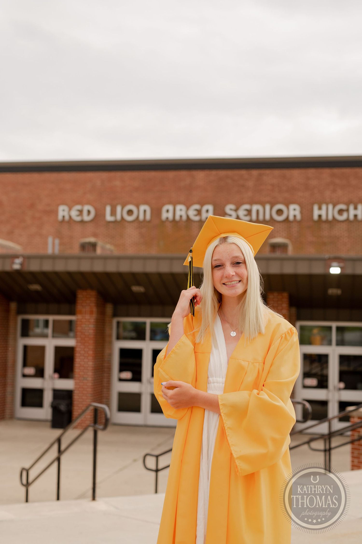 Graduate in yellow graduation attire poses cheerfully outside brick school building.